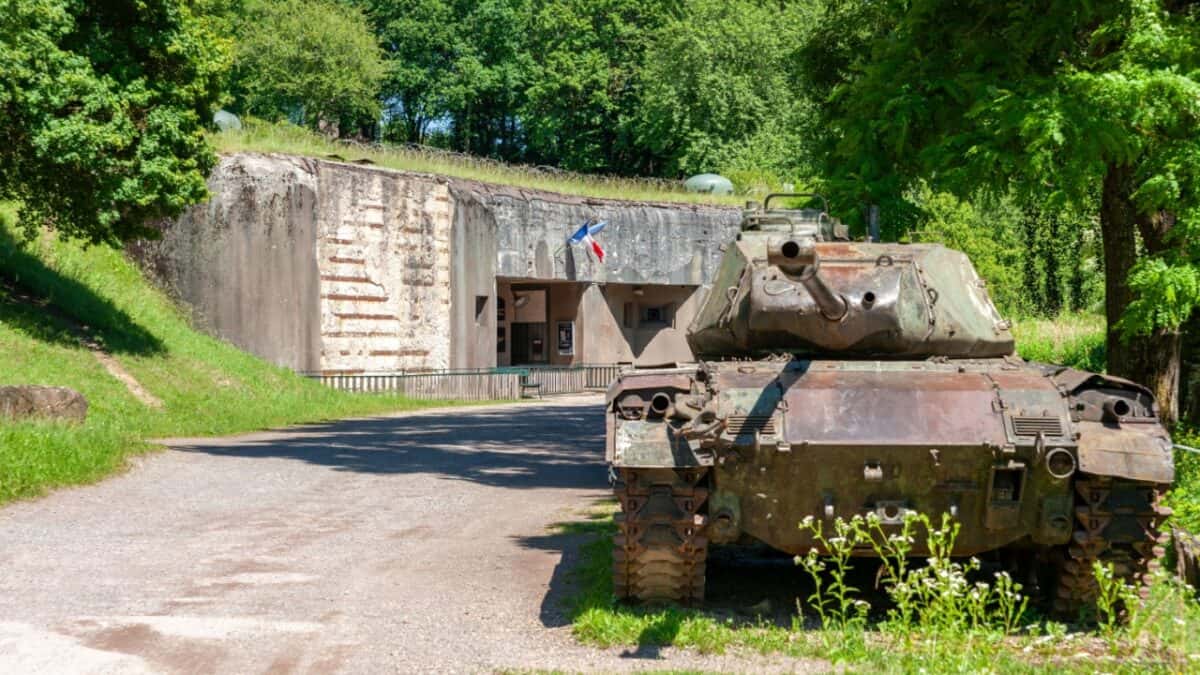 A military tank is parked on a gravel path in front of a concrete bunker entrance, ready for an exciting underground tour. The area is surrounded by lush greenery and trees, with two flags visible above the bunker entrance. This hidden gem makes for a unique travel experience.