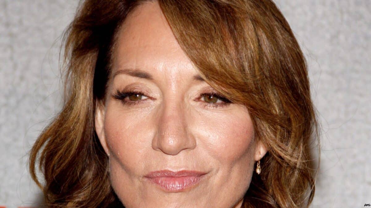 Close-up of a smiling woman with shoulder-length light brown hair and subtle makeup, posing in front of a textured white background.