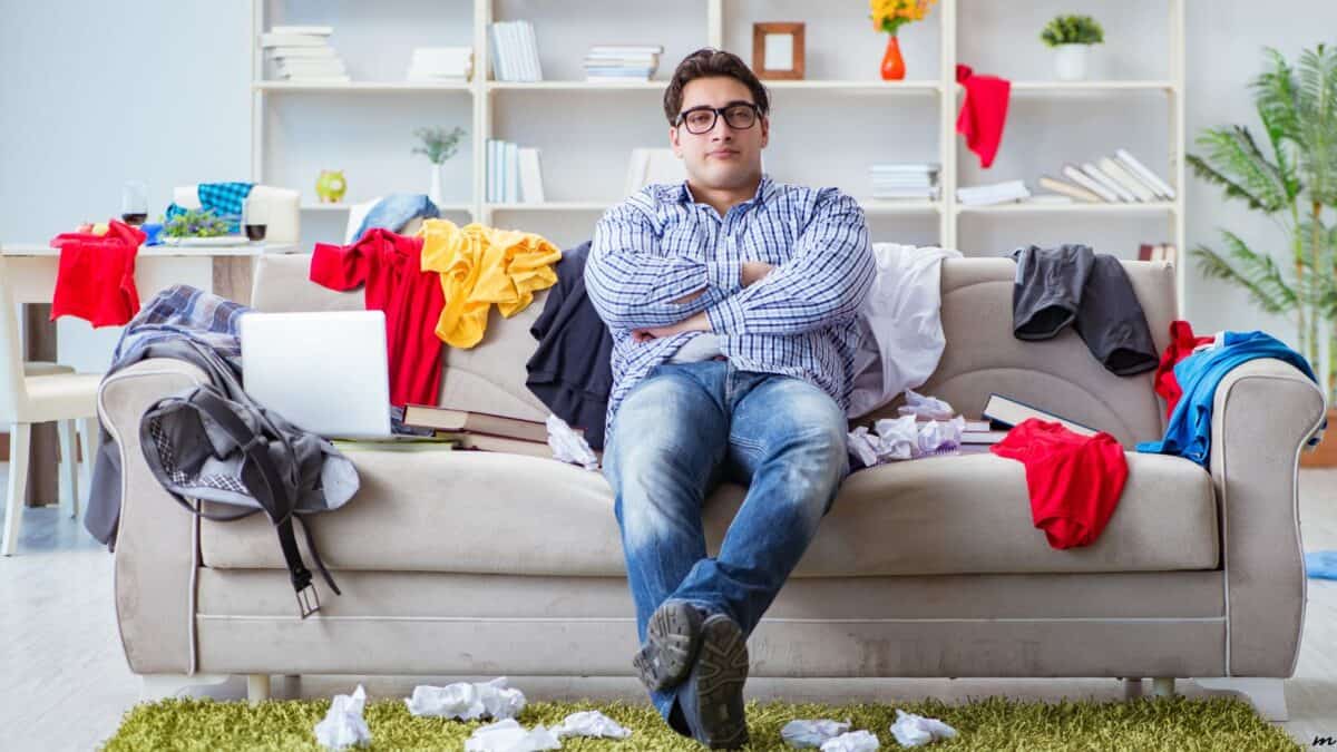 A man sits on a messy couch surrounded by scattered clothes and papers, looking overwhelmed, with a laptop beside him in a cluttered living room.
