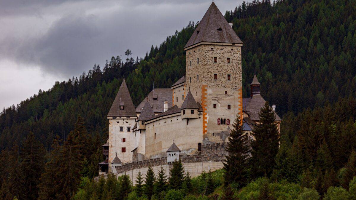 An ancient castle with a prominent stone tower and multiple white walls, nestled among verdant hills and dense green forests under a cloudy sky.