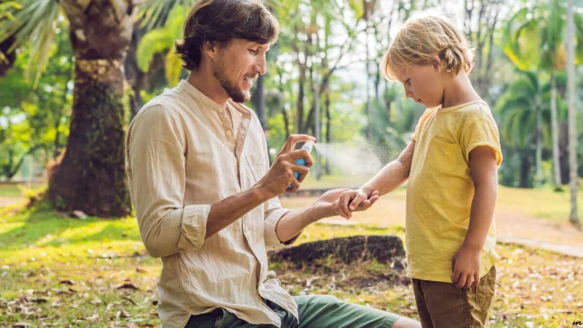 A man applies bug spray to a child's arm in a park with lush green trees.