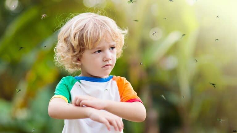 A young child with curly blonde hair swats at mosquitoes outdoors. The child wears a colorful shirt and looks annoyed.