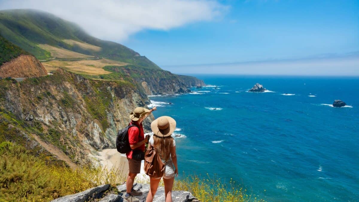 Two people in casual summer attire stand on a cliffside overlooking a scenic coastal view with blue ocean waters, rocky formations, and green hills under a clear, sunny sky. One person has a backpack, and both wear hats. This spot is among the best destinations for emotional recovery.