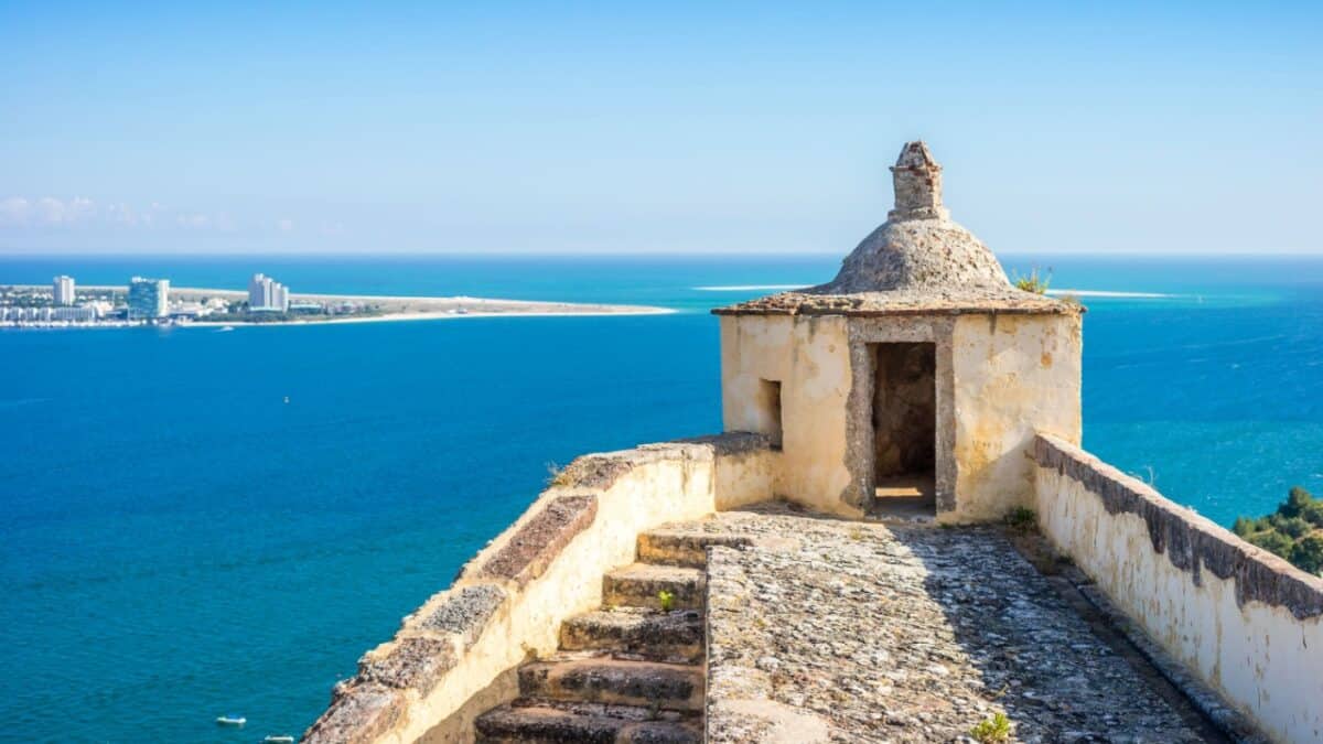 Stone structure with steps on a cliff overlooking a bright blue sea and distant cityscape under a clear sky.