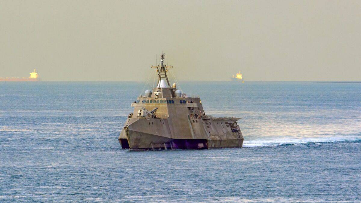 A military stealth warship sailing on a calm sea, with a minimalist, angular design and a radar mast. Two other ships are visible on the horizon against a hazy sky.