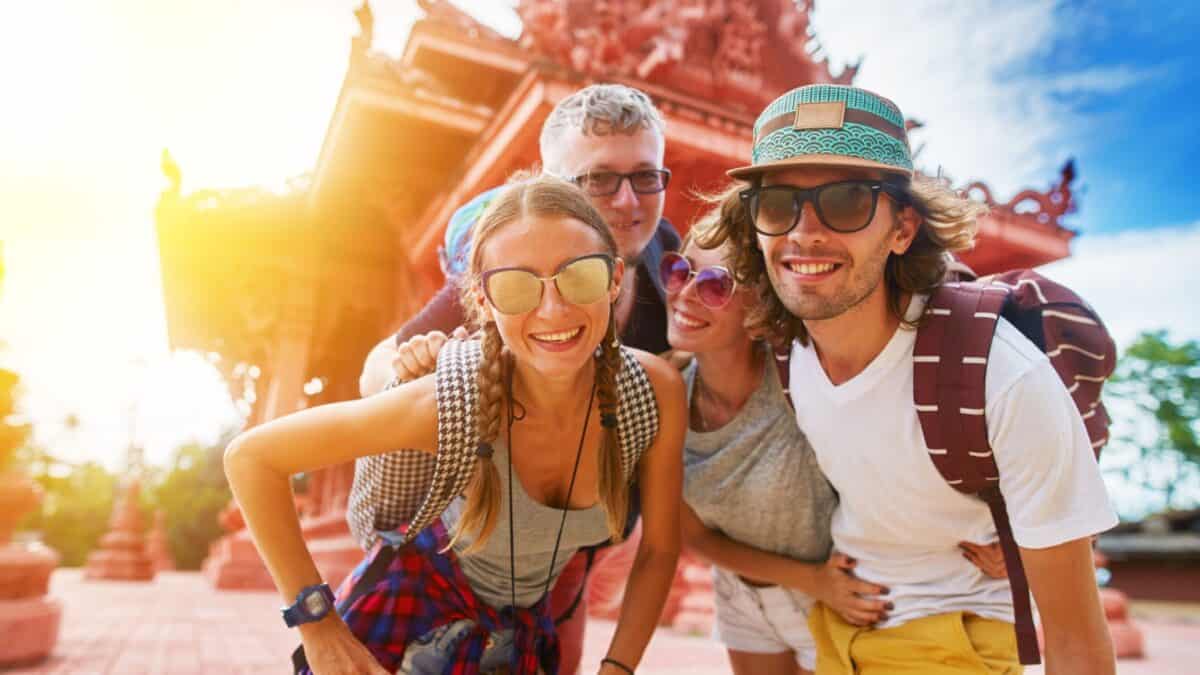 17 Friendliest Cities In The World 17 Four joyful tourists taking a selfie with a brightly lit, ornate temple in the background. They are smiling and wearing casual summer clothes and accessories.
