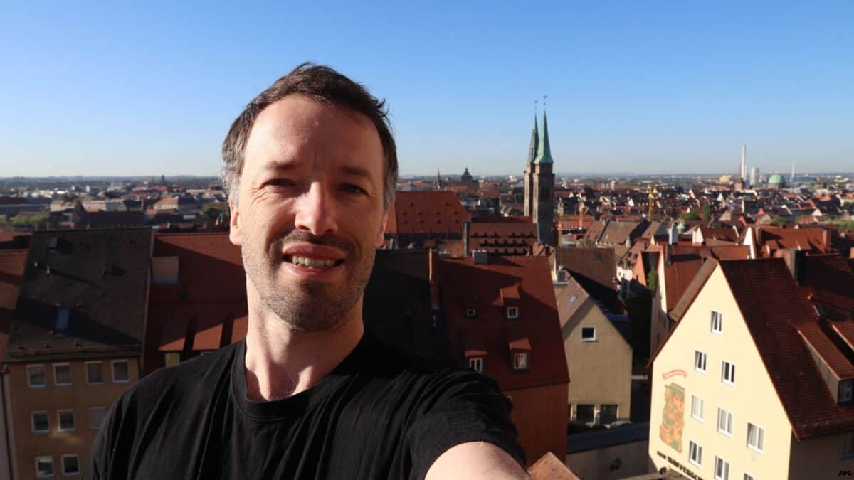 A man smiling in a selfie with a scenic view of a european town's red rooftops and church towers under a clear blue sky in the background.