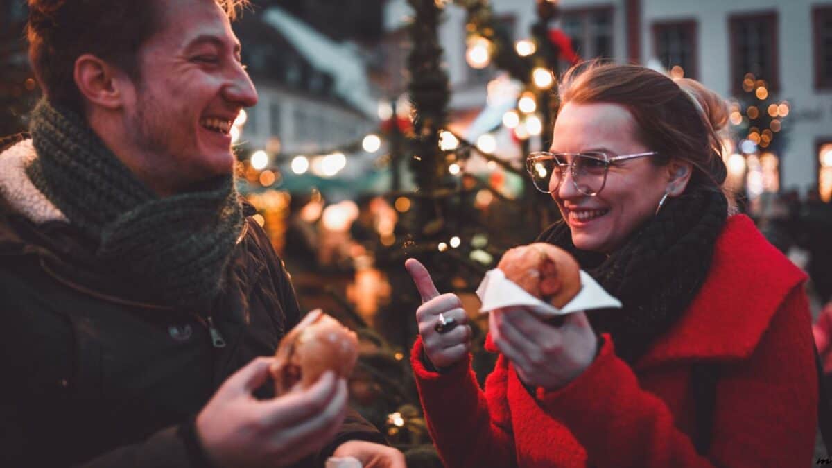 A joyful man and woman, wearing winter clothes, enjoy sausages at a festive outdoor christmas market with lights and a tree in the background.