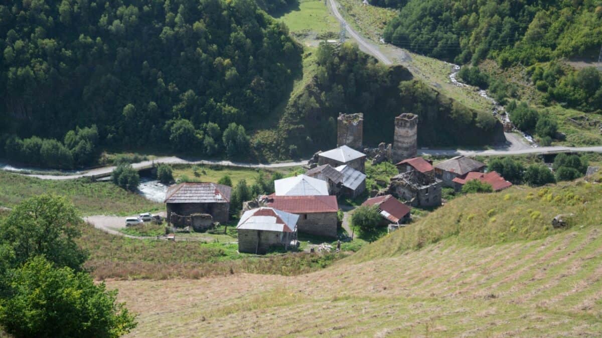 A small ancient village nestled in a green valley surrounded by forested hills. The village consists of a few stone buildings with metal roofs, and two tall, ancient stone towers. A winding road and a river can be seen in the background.