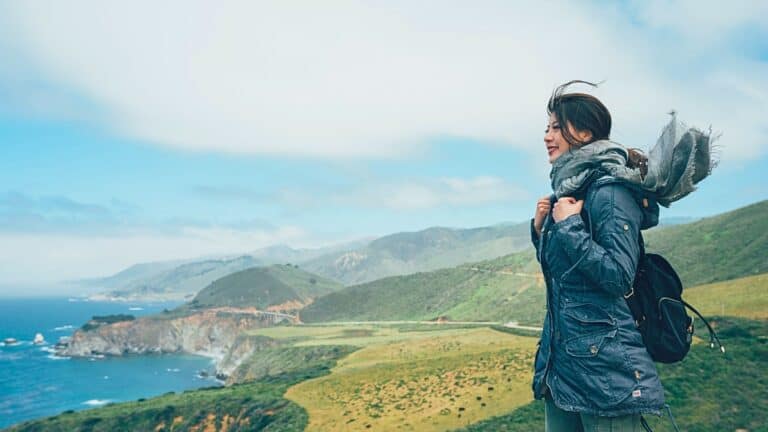 A person wearing a blue jacket and scarf stands on a grassy cliff overlooking the ocean, a scene typical of lesser-known California destinations. The sky is partly cloudy, and rolling hills stretch out in the background. The person is carrying a black backpack and appears to be enjoying the scenic view.