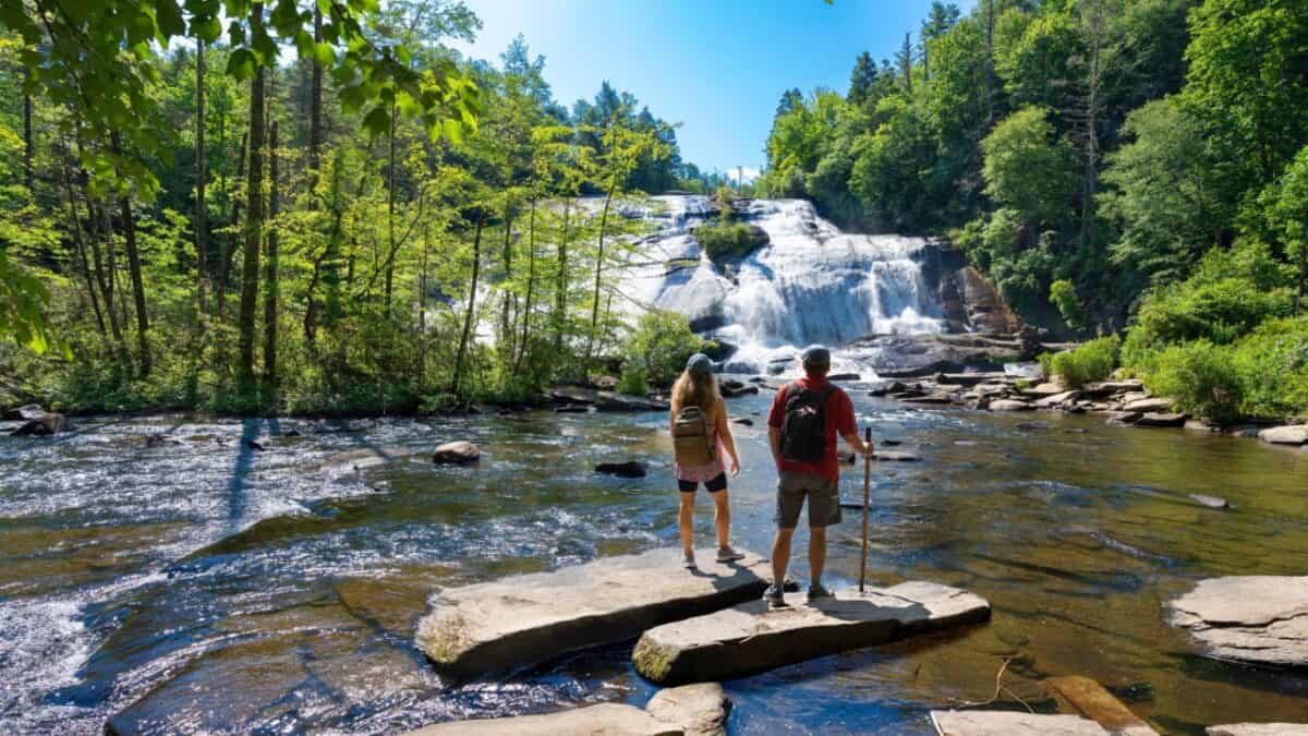 Two outdoor enthusiasts, one in a red shirt and backpack, and the other in a gray shirt, stand on rocks near the edge of a river. They gaze at a large waterfall surrounded by lush green trees under a clear blue sky.