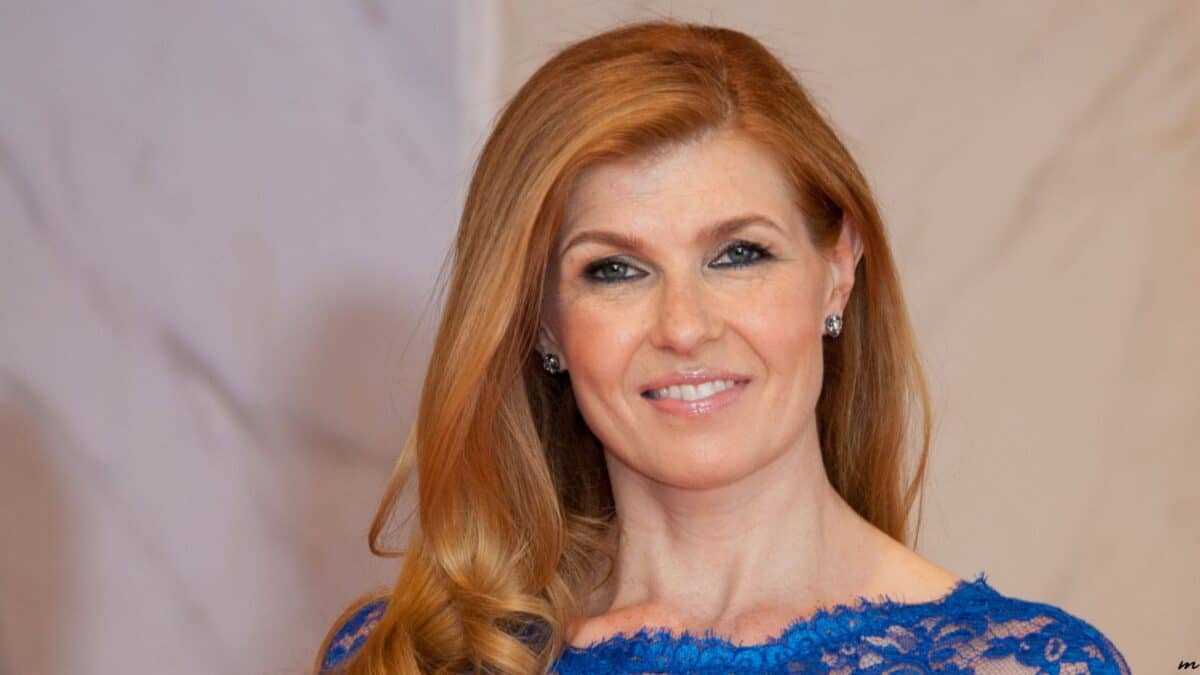 A smiling woman with long red hair wearing a blue lace dress and diamond earrings, standing against a light beige background.