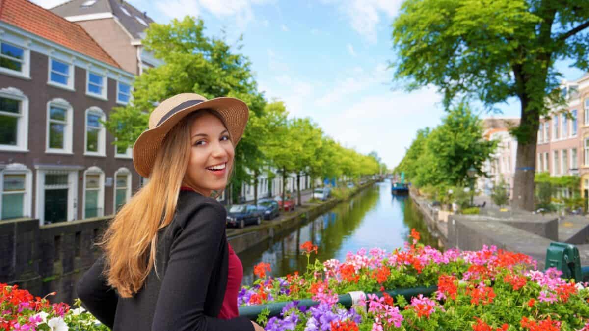 20 Best Places For Solo Female Travelers 20 A woman in a straw hat and black coat stands on a bridge over a canal lined with colorful flowers in a European city. Enjoying her solo travel after breakup, she smiles facing the camera. The canal, flanked by trees and traditional buildings, stretches under a clear blue sky.