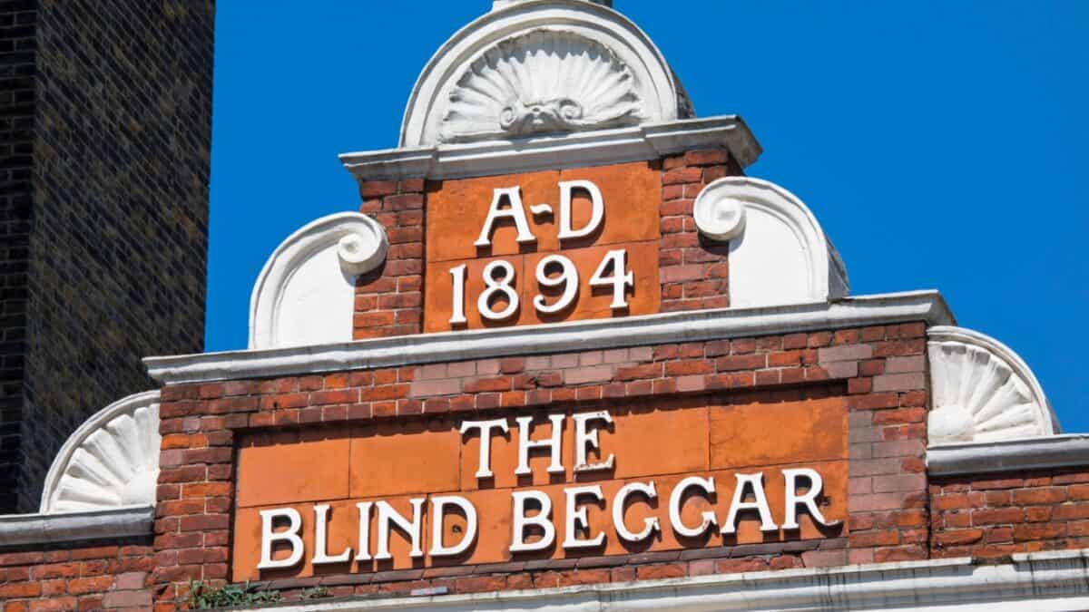 A close-up image of the top section of a brick building with an engraved sign that reads "A.D. 1894" and "THE BLIND BEGGAR" against an orange background. White decorative elements are positioned above and on either side of the sign, evoking the chilling atmosphere often associated with true crime bars.