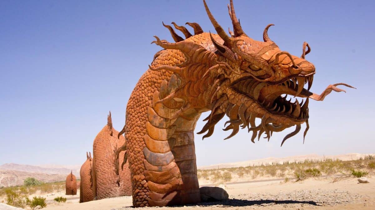 A large, rust-colored metal sculpture of a dragon with intricate scales, claws, and an open mouth stands in a desert landscape. The dragon's body winds through the sandy terrain with a clear blue sky in the background—one of the many lesser-known California destinations worth visiting.