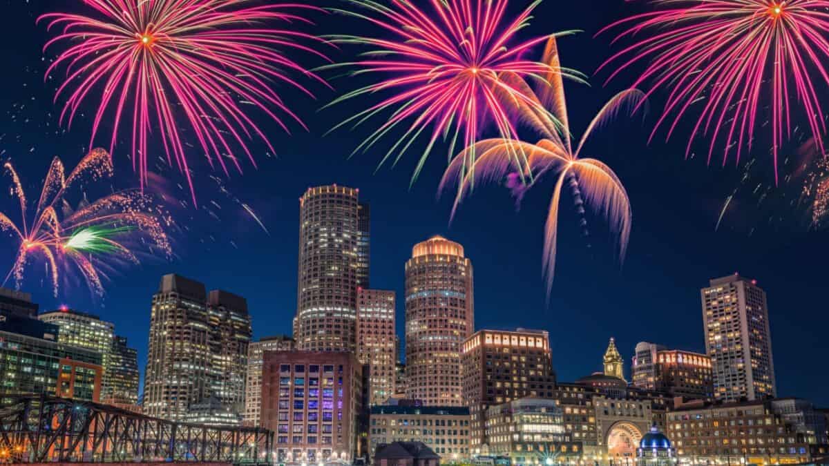 A cityscape at night with a prominent skyline of tall buildings. Bright, colorful fireworks light up the sky in an array of patterns and colors, reflecting off the buildings and creating a festive atmosphere as US cities celebrate the Fourth of July.