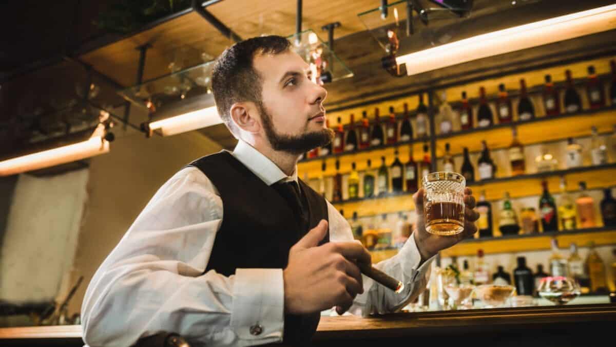 A man with a beard sits at a bar holding a lit cigar in his left hand and a glass of whiskey in his right. Dressed in a white shirt, black vest, and tie, he blends seamlessly into an ambiance reminiscent of True Crime Bars, surrounded by shelves filled with bottles under modern overhead lighting.