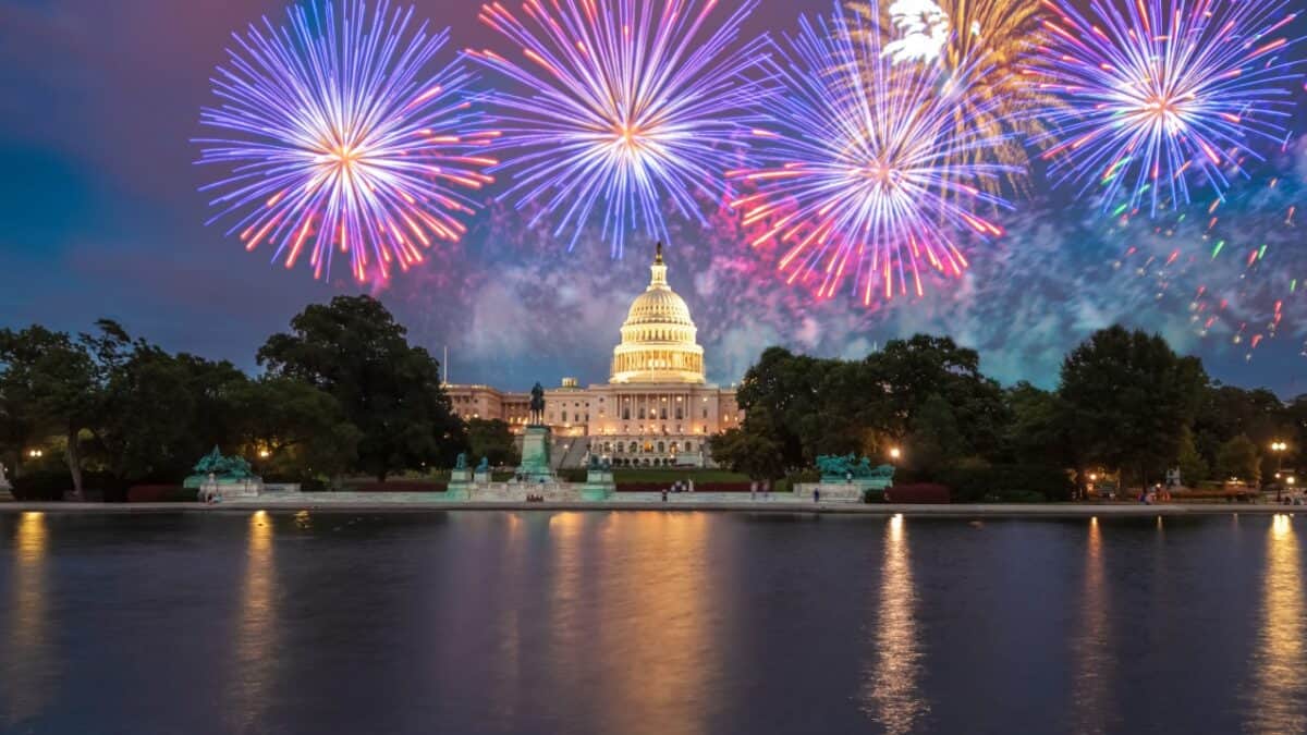The image shows a Fourth of July nighttime scene with colorful fireworks in the sky above the United States Capitol. The Capitol building is illuminated, and its reflection is visible in the water in the foreground, surrounded by trees and lights, capturing a classic celebration in US cities.