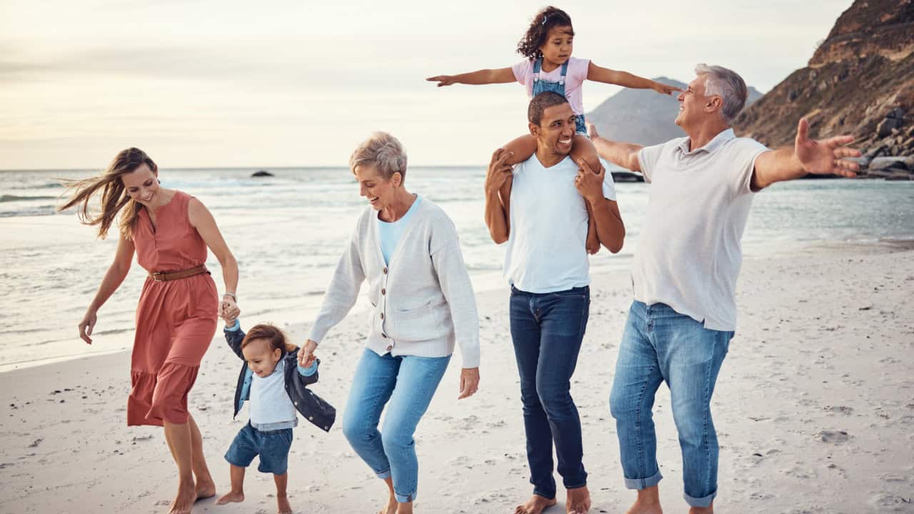 A family of six strolls along a beach, showcasing one of the best locations for family travel. Two children sit atop adults' shoulders as another child holds hands with two more adults. Everyone smiles in their casual clothes, with the stunning ocean and mountains in the background, ideal for multi-generational vacations.