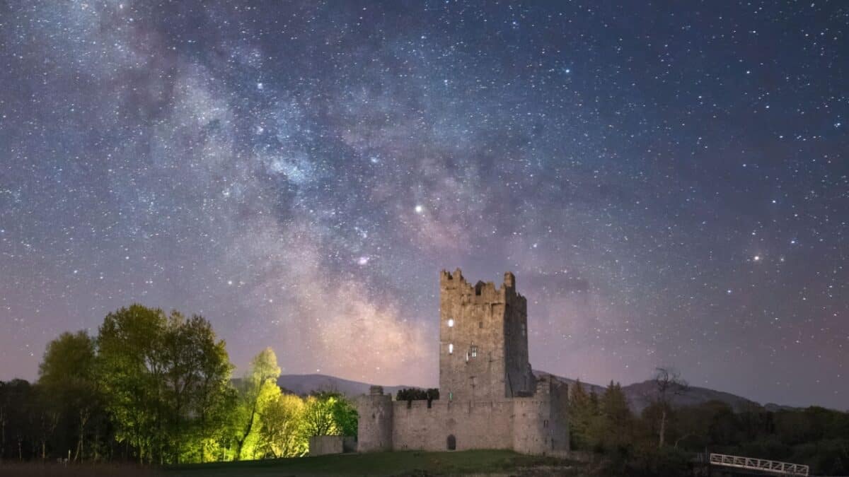 20 Best Places For Solo Female Travelers 8 A stone castle stands illuminated under a star-filled night sky with the Milky Way visible. Trees surround the castle, mountain range in the distance, and a bridge on the right side. One of the secret stargazing spots around the world, it's perfect for those seeking a magical night under the stars.