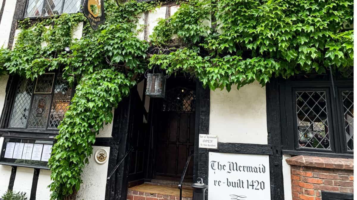 A historic building with ivy-covered walls and diamond-paned windows. A sign reads "The Mermaid re-built 1420." An entrance with a lantern above is flanked by informational signs. This true crime bar, steeped in history and eerie tales, appears to be open for business.