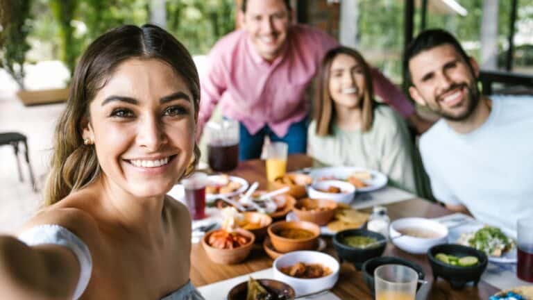 A group of four people are sitting at a table outdoors with a variety of food and drinks in front of them. The person on the left is taking a selfie, and the others are smiling at the camera. They appear to be enjoying a meal together, perhaps brushing up on essential Spanish words while visiting Mexico.