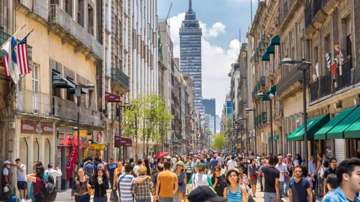 A bustling street scene with numerous people walking along a pedestrianized street lined with old buildings. Some buildings have awnings, and several stores and cafes are visible. In the background, a tall skyscraper stands against a partly cloudy sky—a perfect snapshot for anyone looking to visit Mexico.