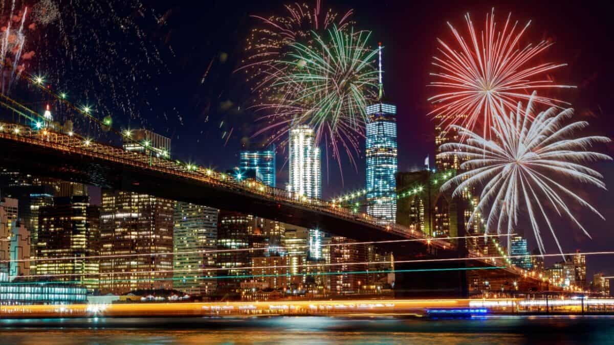 A nighttime cityscape with a large bridge in the foreground and numerous fireworks lighting up the sky, celebrating the Fourth of July. Tall buildings with illuminated windows are visible in the background, and the reflections of the city lights can be seen on the water below.