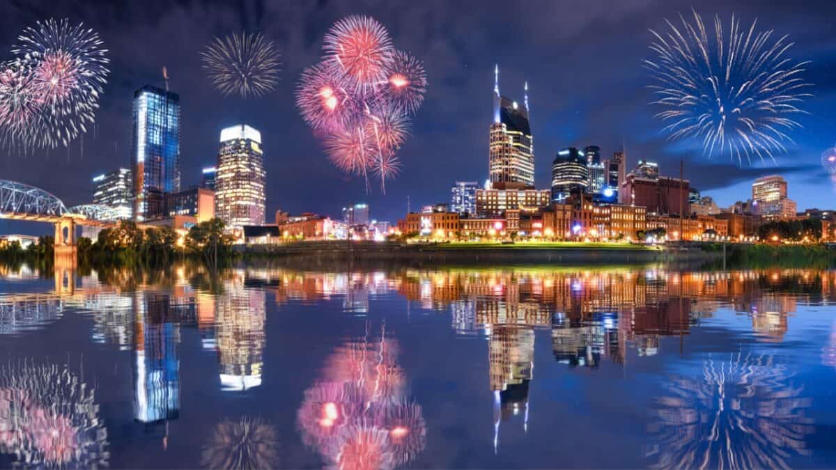 A nighttime cityscape features brightly lit buildings reflecting on a calm body of water. Fireworks of various colors burst in the sky above, celebrating the Fourth of July with vibrant displays. The overall atmosphere is festive and celebratory, capturing the spirit of US cities during this holiday.
