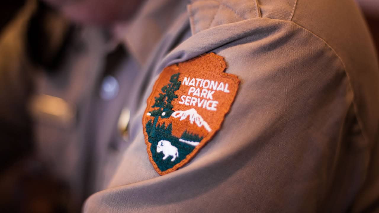 Close-up of a person's arm wearing a National Park Service uniform, showing the embroidered patch with the agency's logo. The patch features a bison, a tree, and mountains with the words "National Park Service" above them, symbolizing their dedication from the grandest to even the smallest national park.