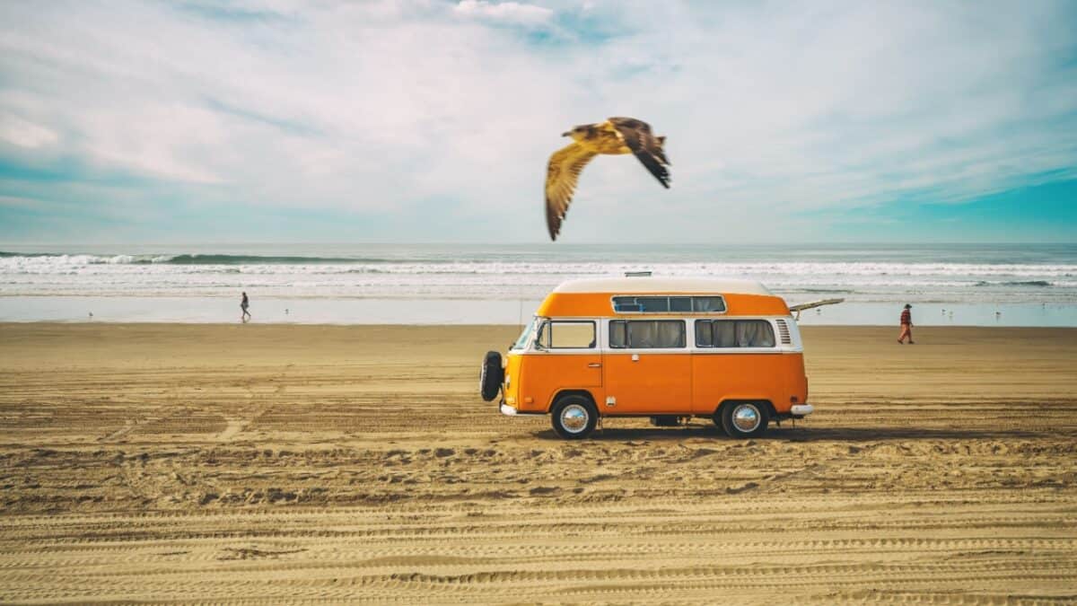 15 Top Travel Destinations for Moving On After a Breakup 5 An orange VW van is parked on a sandy beach in one of the best camping spots California has to offer. The ocean's waves crash in the background, with a bird flying overhead and a person strolling along the water's edge in the distance. The sky is partly cloudy, completing this idyllic scene.