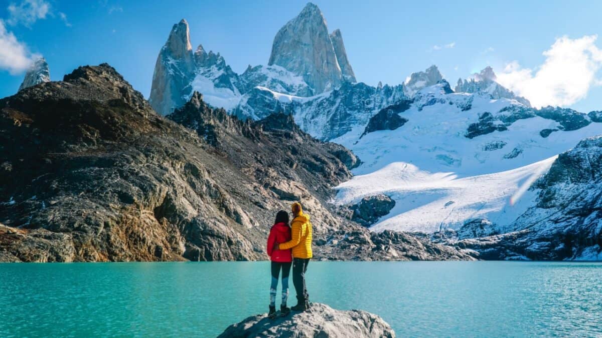 Two people stand on a rock near the edge of a turquoise lake, facing away while looking at a rugged snow-capped mountain landscape on a clear day. One wears a red jacket, the other yellow. This is an ideal visit for adventurous couples seeking unforgettable destinations.