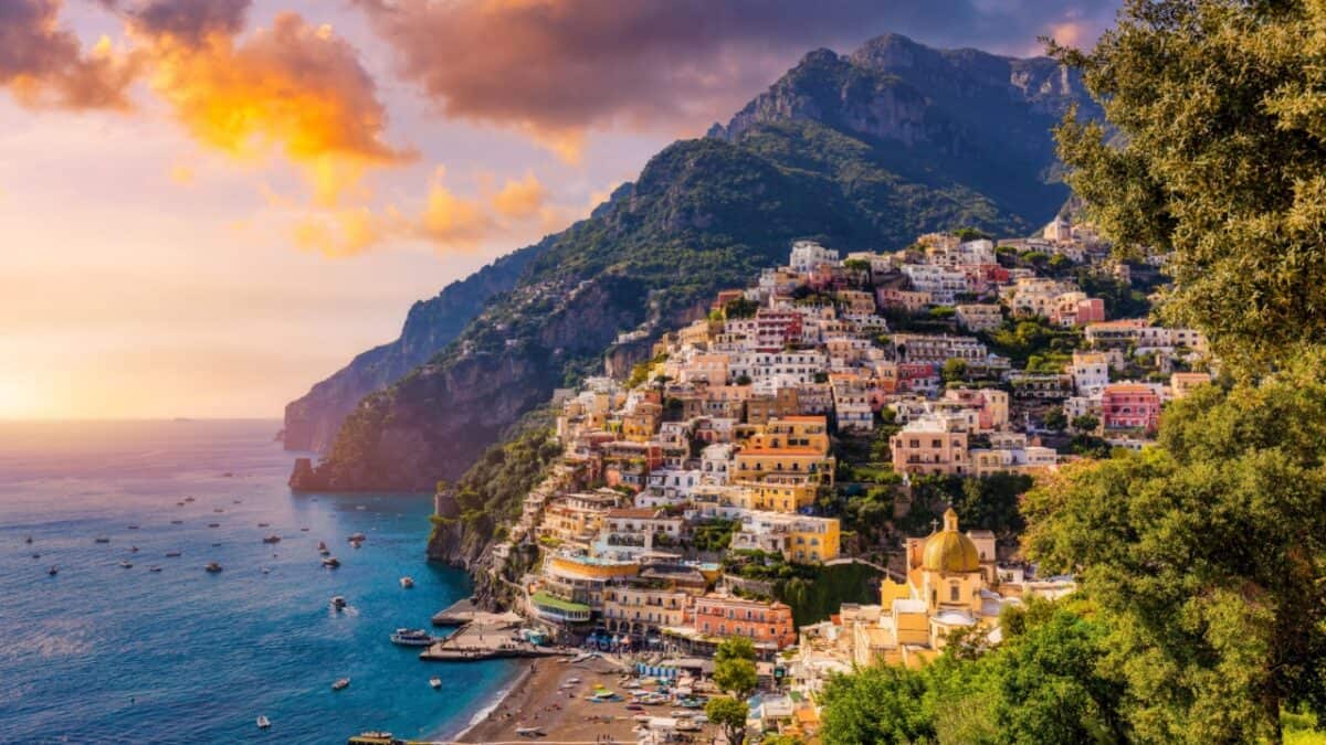 A coastal town with colorful buildings on a hillside overlooking the sea at sunset. The water has many small boats, and the mountains in the background are partially covered by clouds. Trees frame the right side of the image, making it a TikTok Travel hotspot for Viral Travel Locations.