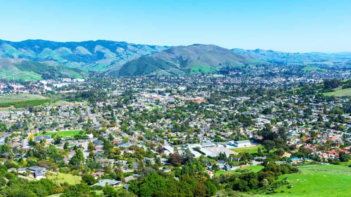 Wide-angle view of a cityscape with a mix of residential and commercial buildings surrounded by green hills and mountains under a clear blue sky. This lesser-known California destination features abundant greenery, a grid-like street layout, and is definitely worth visiting.