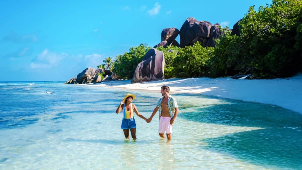 A couple holds hands while walking on a shallow, clear water beach. They are wearing casual summer clothing. In the background are large rock formations, greenery, and a bright blue sky—it's one of those TikTok Travel moments at a Viral Travel Location that you'll want to experience yourself.