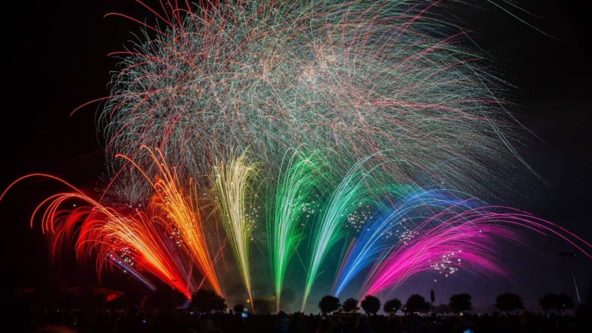 A vibrant fireworks display with multiple colors including red, orange, yellow, green, blue, and pink erupts against a dark night sky. Spectators silhouetted at the bottom watch the spectacle, celebrating the Fourth of July. The fireworks create arcs and bursts of color typical in US cities.