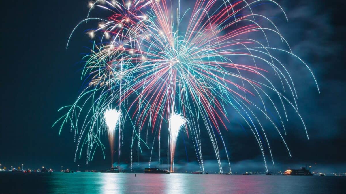 A nighttime scene featuring a Fourth of July fireworks display over water, with bursts of red, blue, green, and white illuminating the sky. Reflections of the fireworks are visible on the water's surface, and distant city lights of US cities can be seen along the horizon as people celebrate.