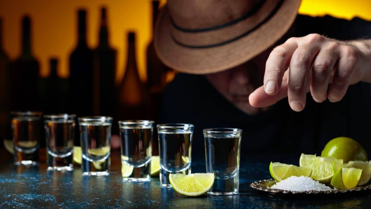 A row of six tequila shots with lime wedges is placed on a counter. A person's hand, wearing a hat, is reaching towards a plate containing lime wedges and salt. It's the kind of scene you'd find in bars everywhere, yet something feels off—as if it belongs in an unsettling true crime story. Bottles are blurred in the background.