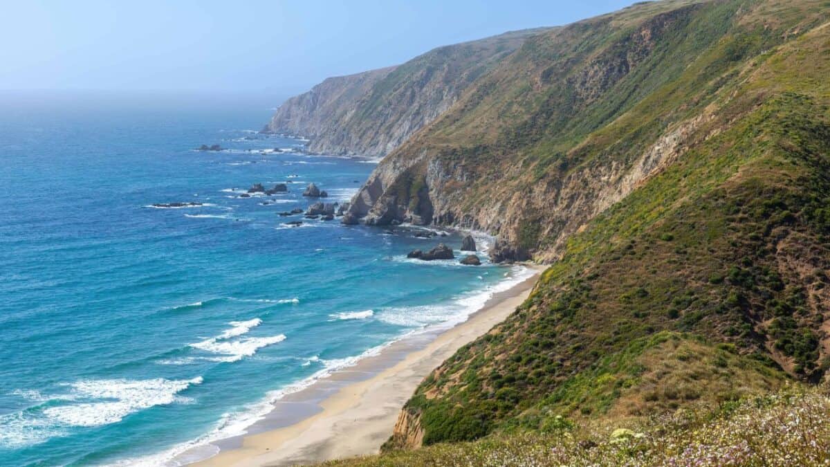 A coastal landscape featuring a sandy beach bordered by turquoise ocean waves on the left, and rocky cliffs with green vegetation on the right. This lesser-known California destination is captured on a clear day with a bright blue sky, and it's definitely worth visiting.