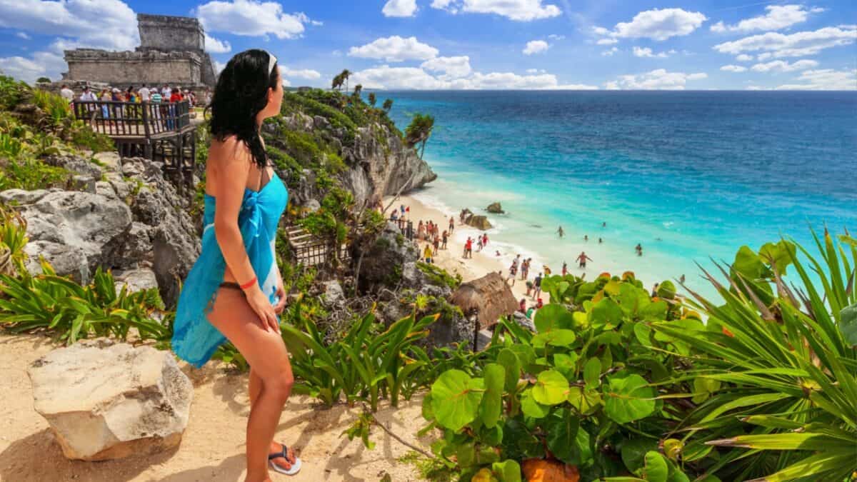 A woman in a blue dress stands on a rocky ledge overlooking a beach with people sunbathing and swimming near turquoise water. There are lush green plants around her and a historical ruin on the cliff in the background, evoking memories of Mexico travel phrases under the scattered blue clouds.