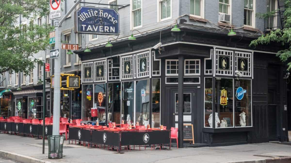Street view of a corner building with a sign reading "White Horse Tavern." The exterior is black with white accents, large windows, and outdoor seating with red chairs and black tables. Neon signs in the windows hint at its bar or pub nature, perfect for those intrigued by true crime bars.
