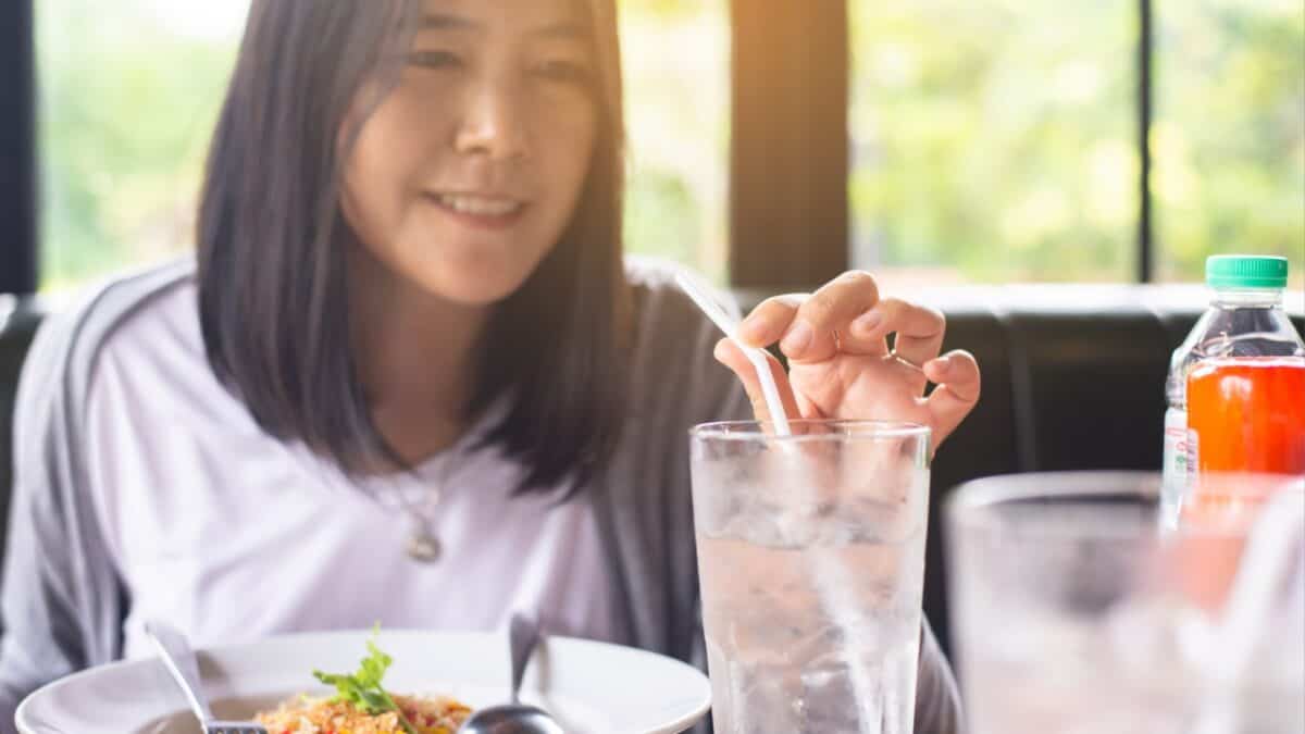 A person with shoulder-length dark hair is sitting at a table in a restaurant. They are holding a bowl of food with their left hand and using their right hand to adjust a straw in a glass of iced water. A bottle with red liquid is also on the table, perhaps reflecting travel etiquette they’ve picked up abroad.