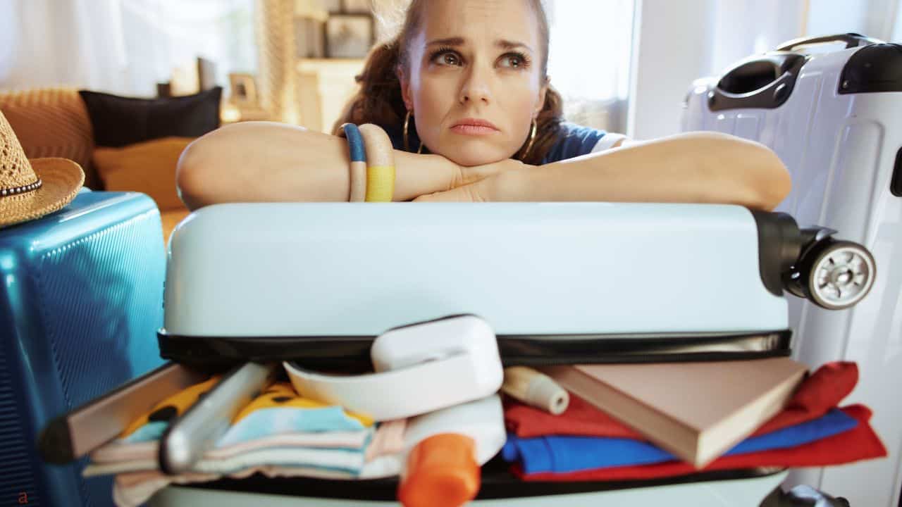 Woman leaning on an overpacked suitcase, looking thoughtful, with travel items spilling out, surrounded by luggage.