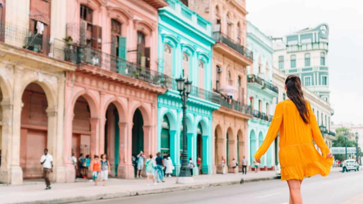 15 Nostalgic Travel Destinations for Anyone Who Grew Up in the '70s 6 A woman in a yellow dress walks down a street lined with colorful, pastel buildings, featuring arches and balconies. Despite the cheerful scene, she considers the safety of female solo travel in unfamiliar destinations. People can be seen in the background.