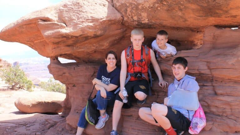 Four people sitting under a rock formation in a desert landscape, smiling at the camera.