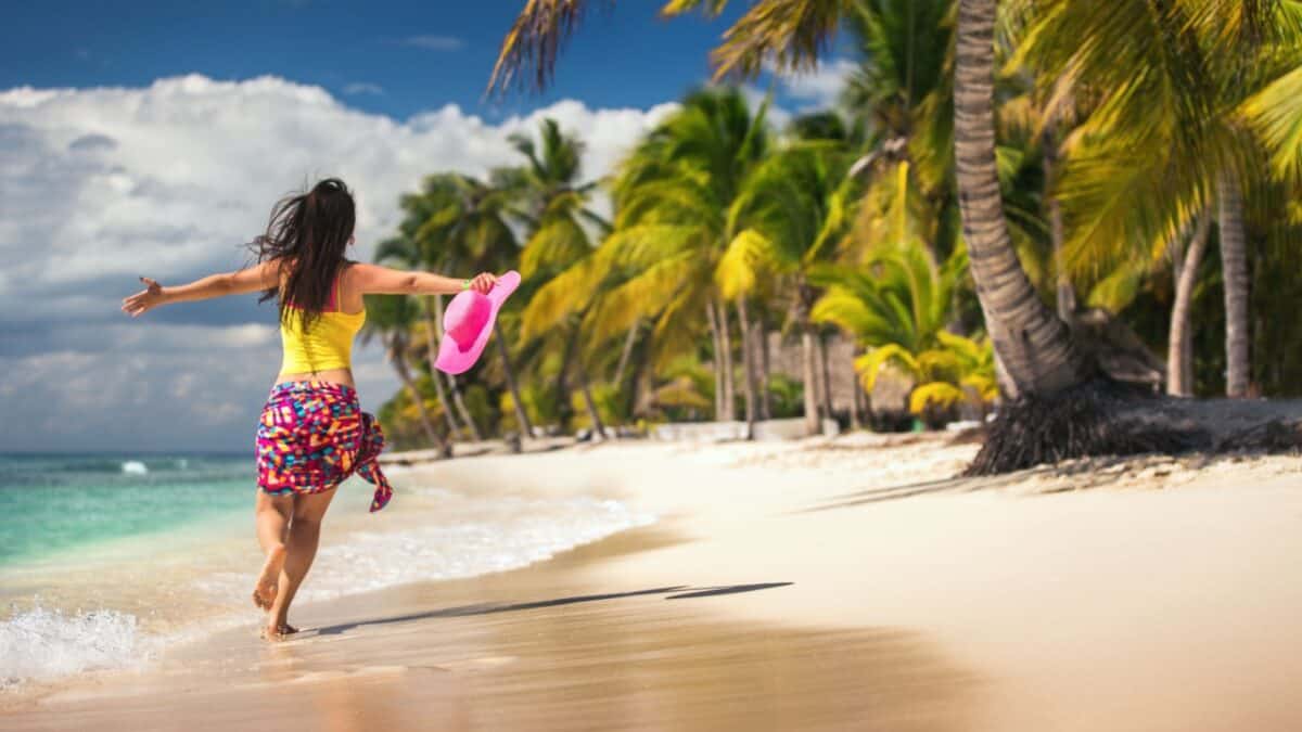 A woman in bright clothing runs along a tropical beach with palm trees, holding a pink hat and spreading her arms out, with the ocean and blue sky in the background