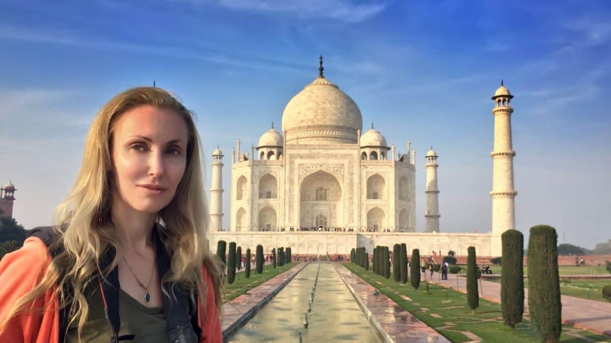 A female solo traveler stands in front of the Taj Mahal, with the iconic white marble mausoleum and its reflecting pool visible in the background on a clear day.