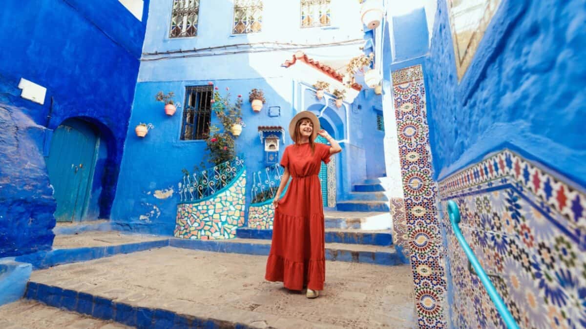 A female solo traveler in a red dress and hat stands on a blue cobbled street with tiled walls, vibrant blue buildings, and colorful decorations, creating a picturesque scene