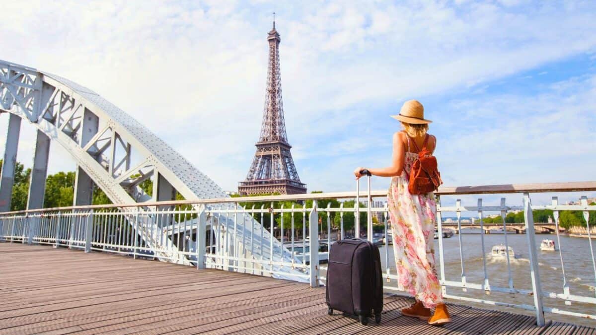 Woman with suitcase and backpack stands on a bridge near the Eiffel Tower, wearing a hat and a floral dress.