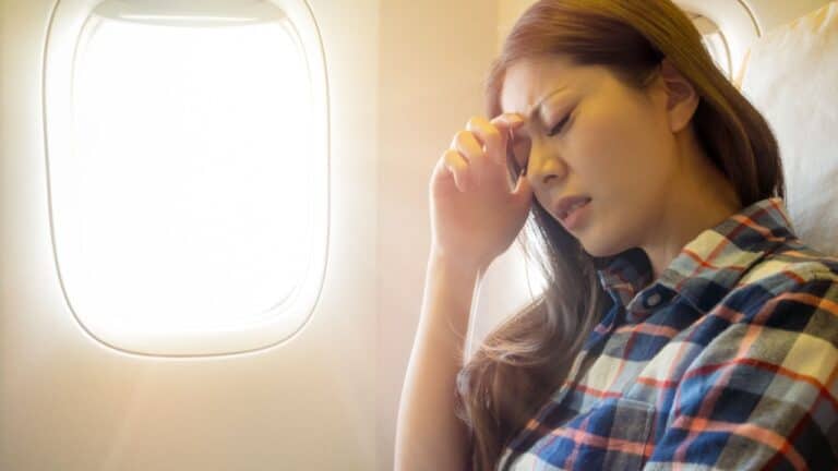 A woman on an airplane looking upset, resting her hand on her forehead while sitting by a window.